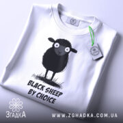 Футболка бути іншим. Біла футболка, складена на столі, з принтом чорної овечки, зображено зверху. Принт чітко видимий, текст "BLACK SHEEP BY CHOICE" розташований під овечкою. На футболці є етикетка з зеленим шнурком.