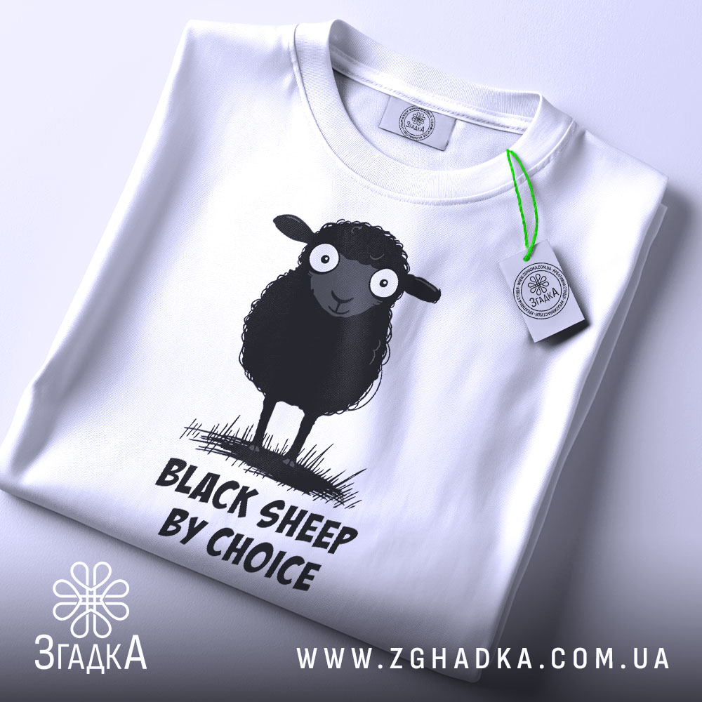 Футболка бути іншим. Біла футболка, складена на столі, з принтом чорної овечки, зображено зверху. Принт чітко видимий, текст "BLACK SHEEP BY CHOICE" розташований під овечкою. На футболці є етикетка з зеленим шнурком.