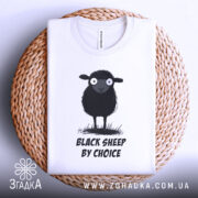 Футболка бути іншим. Біла футболка, складена на плетеному кошику, з принтом чорної овечки, зображено зверху. Принт чітко видимий, текст "BLACK SHEEP BY CHOICE" розташований під овечкою. Кошик має натуральний колір, що контрастує з футболкою.