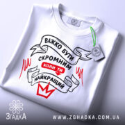 Футболка бути скромним, біла, складена на столі, фон світло-сірий, принт чорного та червоного кольорів з написом 'ВАЖКО БУТИ СКРОМНИМ КОЛИ ТИ НАЙКРАЩИЙ', з зеленим ярликом.