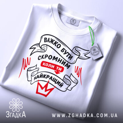 Футболка бути скромним, біла, складена на столі, фон світло-сірий, принт чорного та червоного кольорів з написом 'ВАЖКО БУТИ СКРОМНИМ КОЛИ ТИ НАЙКРАЩИЙ', з зеленим ярликом.