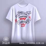 Футболка бути скромним, біла, зображена на вішалці, фон світло-сірий, принт чорного та червоного кольорів з написом 'ВАЖКО БУТИ СКРОМНИМ КОЛИ ТИ НАЙКРАЩИЙ'.