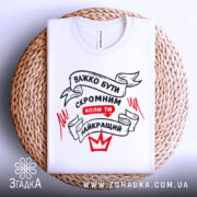 Футболка бути скромним, біла, складена на плетеному кошику, фон білий, принт чорного та червоного кольорів з написом 'ВАЖКО БУТИ СКРОМНИМ КОЛИ ТИ НАЙКРАЩИЙ'.