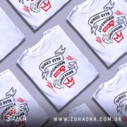 Футболка бути скромним, біла, зображена на манекені, фон світло-сірий, принт чорного та червоного кольорів з написом 'ВАЖКО БУТИ СКРОМНИМ КОЛИ ТИ НАЙКРАЩИЙ'.