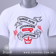 Футболка бути скромним, біла, складена на столі, фон світло-сірий, принт чорного та червоного кольорів з написом 'ВАЖКО БУТИ СКРОМНИМ КОЛИ ТИ НАЙКРАЩИЙ', з зеленим ярликом.