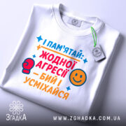 Футболка бий і усміхайся. Біла футболка, складена на столі, з кольоровим принтом, що містить текст та малюнки. На футболці є бірка з логотипом. Фон світло-сірий.