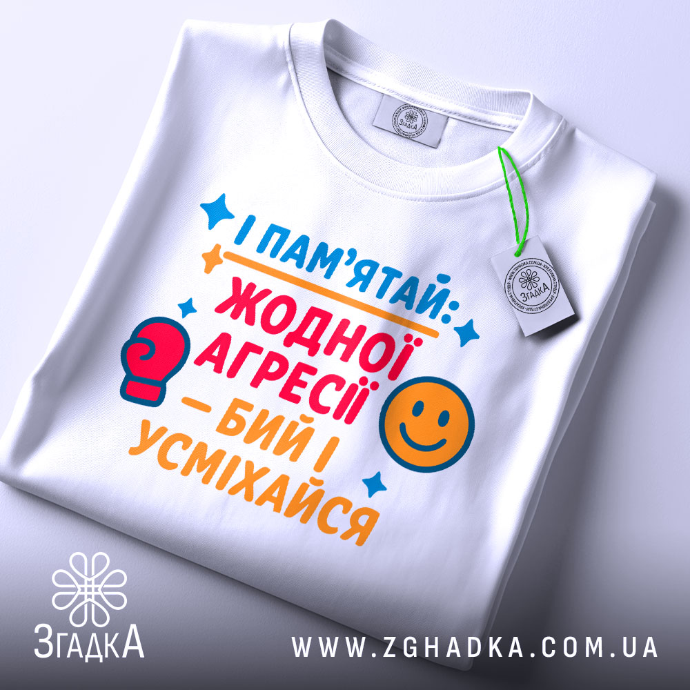 Футболка бий і усміхайся. Біла футболка, складена на столі, з кольоровим принтом, що містить текст та малюнки. На футболці є бірка з логотипом. Фон світло-сірий.