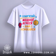 Футболка бий і усміхайся. Біла футболка, підвішена на вішалці, з яскравим кольоровим принтом на грудях. Текст на футболці червоний, жовтий та синій. Фон світло-сірий.