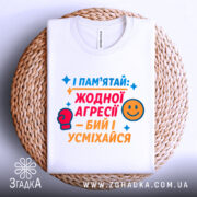 Футболка бий і усміхайся. Біла футболка, складена на плетеній кошику, з кольоровим принтом, що містить текст та малюнки. Фон світло-сірий.