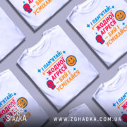 Футболка бий і усміхайся. Біла футболка, складена на столі, з кольоровим принтом, що містить текст та малюнки. Фон світло-сірий.