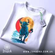 Футболка бик енергія, біла, складена на поверхні, фон світло-сірий, принт з биком, що біжить на фоні червоного сонця, з етикеткою на шнурку.