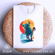 Футболка бик енергія, біла, складена на плетеній кошику, фон світло-сірий, принт з биком, що біжить на фоні червоного сонця, видно етикетку на комірці.