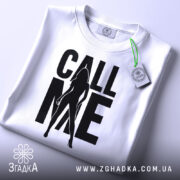 Футболка call me білого кольору, складена на столі, фон світло-сірий, принт чорного кольору з силуетом жінки та написом 'CALL ME', на комірці видна етикетка.