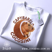 Футболка Capybarelo Cocosini білого кольору, зображена зверху, на ній видно етикетку з зеленим шнурком, фон світло-сірий, принт з капібарою в кокосі яскравий і чіткий.