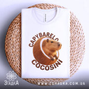 Футболка Capybarelo Cocosini білого кольору, зображена зверху на плетеній кошику, фон світло-сірий, принт з капібарою в кокосі чіткий і яскравий, видна етикетка.