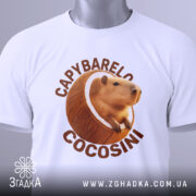Футболка Capybarelo Cocosini білого кольору, зображена зверху, на ній видно етикетку з зеленим шнурком, фон світло-сірий, принт з капібарою в кокосі яскравий і чіткий.