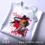 Футболка Chainsaw Man білого кольору, складена на поверхні, зображена з верхнього ракурсу, фон світлий, принт з персонажем з бензопилою, оранжевий шолом, червоні та чорні плями навколо.