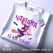 Футболка чарівна на голову, біла, показана з верхнього ракурсу, на ній є етикетка з зеленим шнурком, фон світлий, принт з чарівницею на мітлі та летючими кажанами, текст 'Чарівна на всю голову' в фіолетовому кольорі.