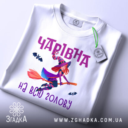 Футболка чарівна на голову, біла, показана з верхнього ракурсу, на ній є етикетка з зеленим шнурком, фон світлий, принт з чарівницею на мітлі та летючими кажанами, текст 'Чарівна на всю голову' в фіолетовому кольорі.
