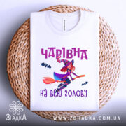 Футболка чарівна на голову, біла, показана зверху на плетеній основі, фон світлий, принт з чарівницею на мітлі та летючими кажанами, текст 'Чарівна на всю голову' в фіолетовому кольорі.