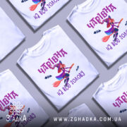 Футболка чарівна на голову, біла, показана з фронтального ракурсу на манекені, фон нейтральний, принт з зображенням чарівниці на мітлі з червоним волоссям і летючими кажанами, текст 'Чарівна на всю голову' в фіолетовому кольорі.