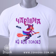 Футболка чарівна на голову, біла, показана з верхнього ракурсу, на ній є етикетка з зеленим шнурком, фон світлий, принт з чарівницею на мітлі та летючими кажанами, текст 'Чарівна на всю голову' в фіолетовому кольорі.