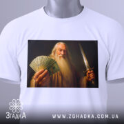 Футболка чарівник з доларами, біла, складена на столі, фон білий, принт зображає чоловіка з довгим волоссям і бородою, який тримає гроші і меч, на футболці є етикетка з зеленим шнурком.