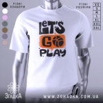 Футболка час грати баскетбол, біла футболка на манекені, з принтом у вигляді напису 'LET'S GO PLAY' з оранжевим фоном та чорними літерами, фон світло-сірий, деталі принта чітко видимі.