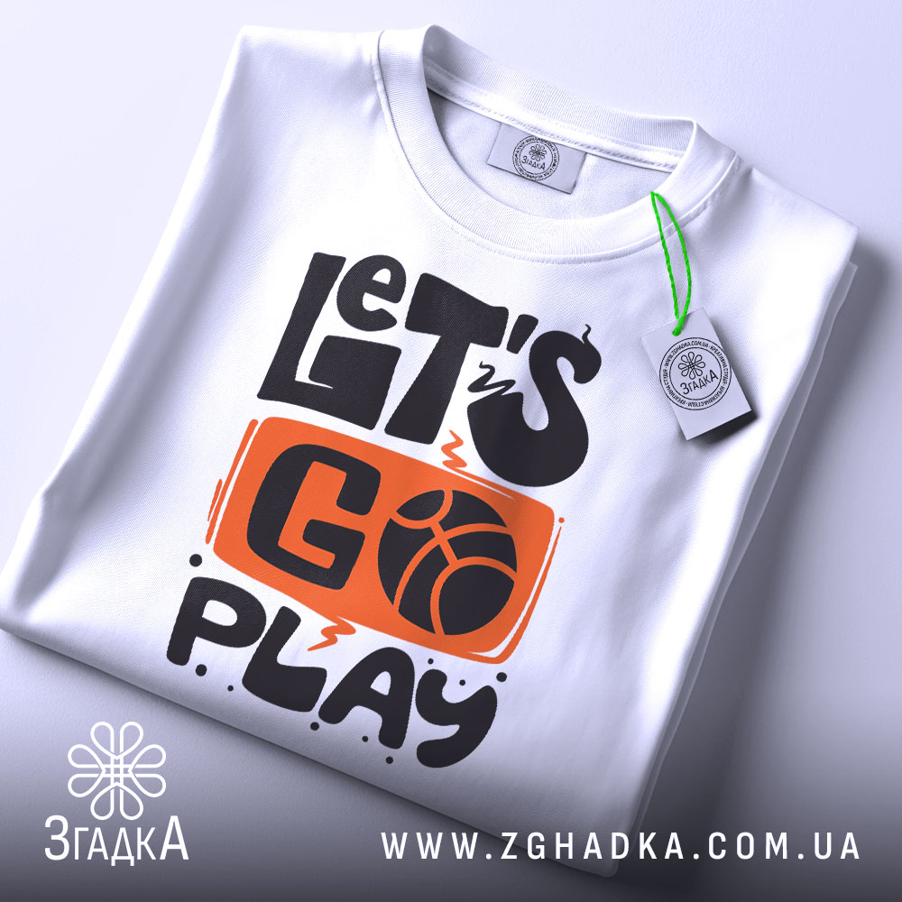 Футболка час грати баскетбол, біла футболка, складена на поверхні, з принтом 'LET'S GO PLAY', оранжевий фон з чорними літерами, на комірці є етикетка, фон світло-сірий.