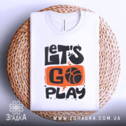 Футболка час грати баскетбол, біла футболка, складена на плетеній кошику, з принтом 'LET'S GO PLAY' в оранжевому та чорному кольорах, фон світло-сірий, футболка виглядає акуратно.