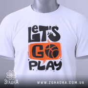 Футболка час грати баскетбол, біла футболка, складена на поверхні, з принтом 'LET'S GO PLAY', оранжевий фон з чорними літерами, на комірці є етикетка, фон світло-сірий.