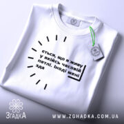 Футболка часова петля, біла, складена на поверхні, фон світло-сірий, принт чорного кольору з текстом, на комірці є етикетка з зеленим шнурком.