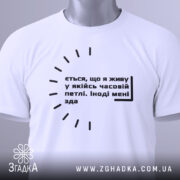 Футболка часова петля, біла, складена на поверхні, фон світло-сірий, принт чорного кольору з текстом, на комірці є етикетка з зеленим шнурком.