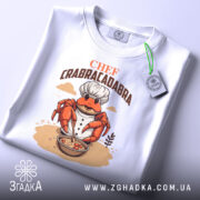 Футболка Chef Crabracadabra білого кольору, зображена в складеному вигляді, фон світло-сірий, принт з крабом у кухарському костюмі, який готує страву в мисці, на етикетці зелена стрічка.