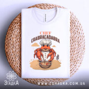 Футболка Chef Crabracadabra білого кольору, зображена в складеному вигляді на плетеній кошику, фон світло-сірий, принт з крабом у кухарському костюмі, який готує страву в мисці, акцент на текстурі кошика.