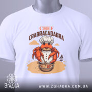 Футболка Chef Crabracadabra білого кольору, зображена в складеному вигляді, фон світло-сірий, принт з крабом у кухарському костюмі, який готує страву в мисці, на етикетці зелена стрічка.