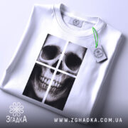 Футболка череп модний принт, біла, складена на поверхні, фон світло-сірий, принт зображає череп у чотирьох квадратах, на футболці є етикетка з зеленим шнурком.
