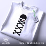 Футболка череп символ, біла, зображена зверху, складена на столі, фон світлий, принт чорного черепа з червоною краплею та кістками, з етикеткою.