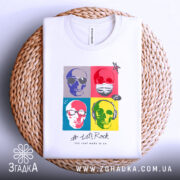 Футболка черепи рок стиль, біла, розкладена на плетеній кошику, фон світло-сірий, принт з чотирьох черепів у кольорових квадратах, текст '# Let's Rock' під принтом, видна етикетка на комірці.