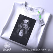 Футболка черниця тату білого кольору, складена на поверхні з легким відблиском, фон світло-сірий, принт з чорно-білою фотографією дівчини в чернечому вбранні, на футболці є етикетка з зеленим шнурком.