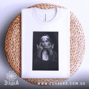 Футболка черниця тату білого кольору, складена на плетеній кошику, фон світло-сірий, принт з чорно-білою фотографією дівчини в чернечому вбранні, видно етикетку з написом.