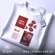 Футболка червоний акцент, біла футболка з принтом, зображена зверху, фон світлий, принт містить червоні губи та зображення червоного автомобіля, на футболці є етикетка.