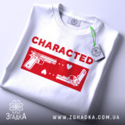 Футболка червоний стиль, складена біла футболка з принтом червоного кольору, на якому зображені два пістолети та напис 'CHARACTED', фон світло-сірий, видно етикетку з зеленим шнурком.