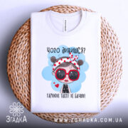 Футболка чого дивишся? Біла футболка, складена на плетеній основі, з принтом дівчинки в червоних окулярах, фон світло-сірий, принт яскравий, видно етикетку.