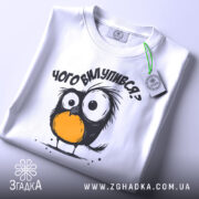 Футболка чого вилупився, білого кольору, складена на поверхні, фон світлий, принт з пташеням з великими очима та оранжевим животом, на футболці є етикетка.