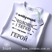 Футболка чоловік і татко, біла, складена на плоскій поверхні, фон світло-сірий, принт чорного кольору з написом 'найкращий чоловік і татко ми наш герой', з зеленим ярликом.