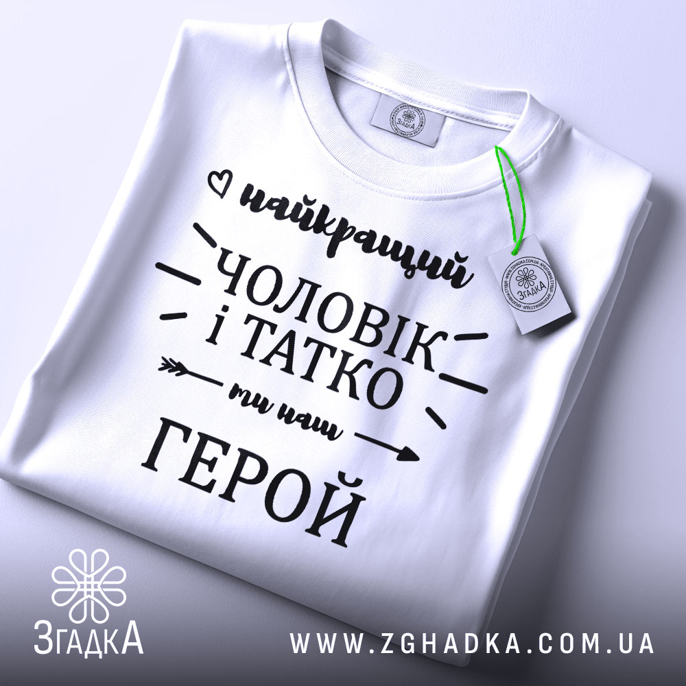 Футболка чоловік і татко, біла, складена на плоскій поверхні, фон світло-сірий, принт чорного кольору з написом 'найкращий чоловік і татко ми наш герой', з зеленим ярликом.