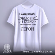 Футболка чоловік і татко, біла, зображена з фронтального ракурсу на вішалці, фон світло-сірий, принт чорного кольору з написом 'найкращий чоловік і татко ми наш герой'.