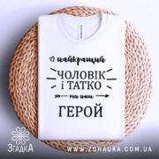 Футболка чоловік і татко, біла, складена на плоскій поверхні з плетеного кошика, фон світло-сірий, принт чорного кольору з написом 'найкращий чоловік і татко ми наш герой'.