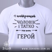 Футболка чоловік і татко, біла, складена на плоскій поверхні, фон світло-сірий, принт чорного кольору з написом 'найкращий чоловік і татко ми наш герой', з зеленим ярликом.